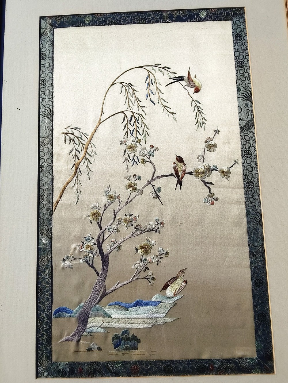 A framed Chinese silk embroidery picture depicting birds perched among blossoming branches and willow leaves on a neutral cream background with a decorative blue brocade border.