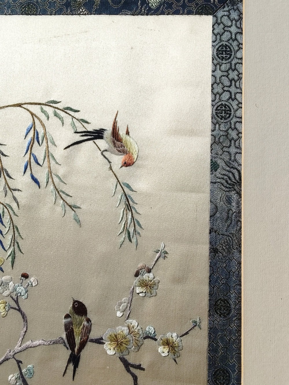 A framed Chinese silk embroidery picture depicting birds perched among blossoming branches and willow leaves on a neutral cream background with a decorative blue brocade border. View II