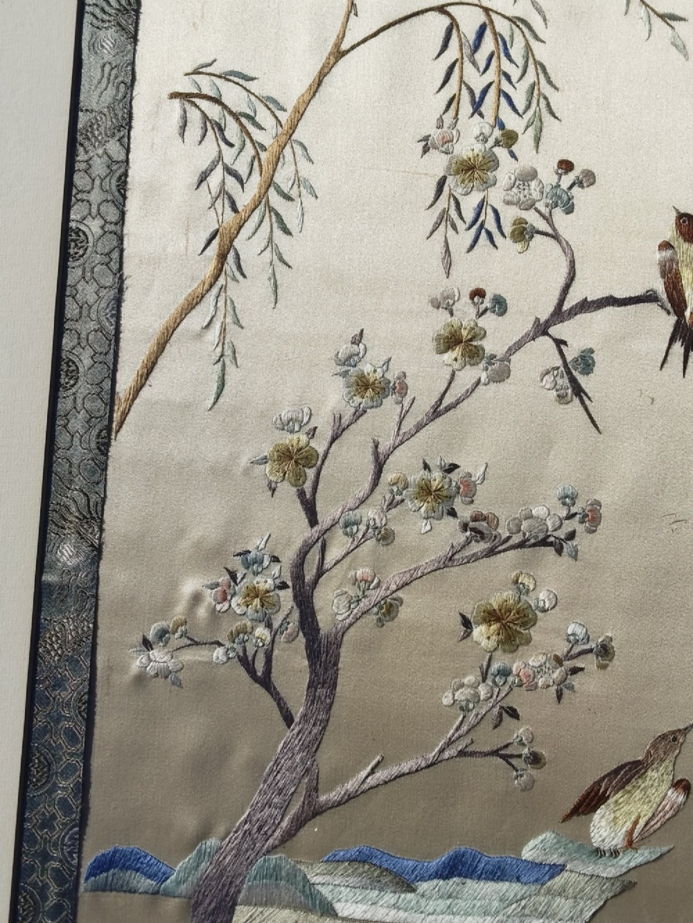A framed Chinese silk embroidery picture depicting birds perched among blossoming branches and willow leaves on a neutral cream background with a decorative blue brocade border. View IV