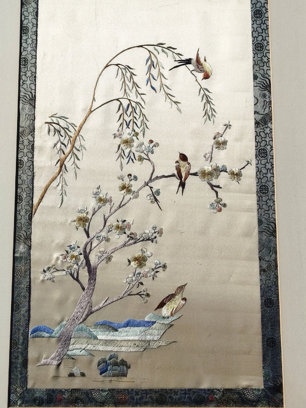 A framed Chinese silk embroidery picture depicting birds perched among blossoming branches and willow leaves on a neutral cream background with a decorative blue brocade border. View V