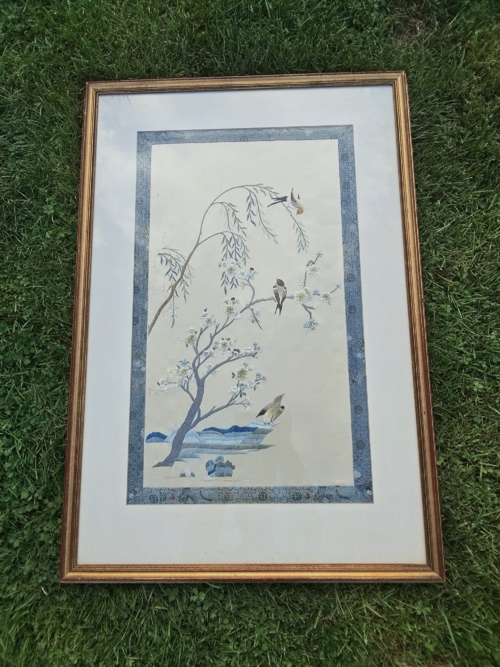 A framed Chinese silk embroidery picture depicting birds perched among blossoming branches and willow leaves on a neutral cream background with a decorative blue brocade border. View X