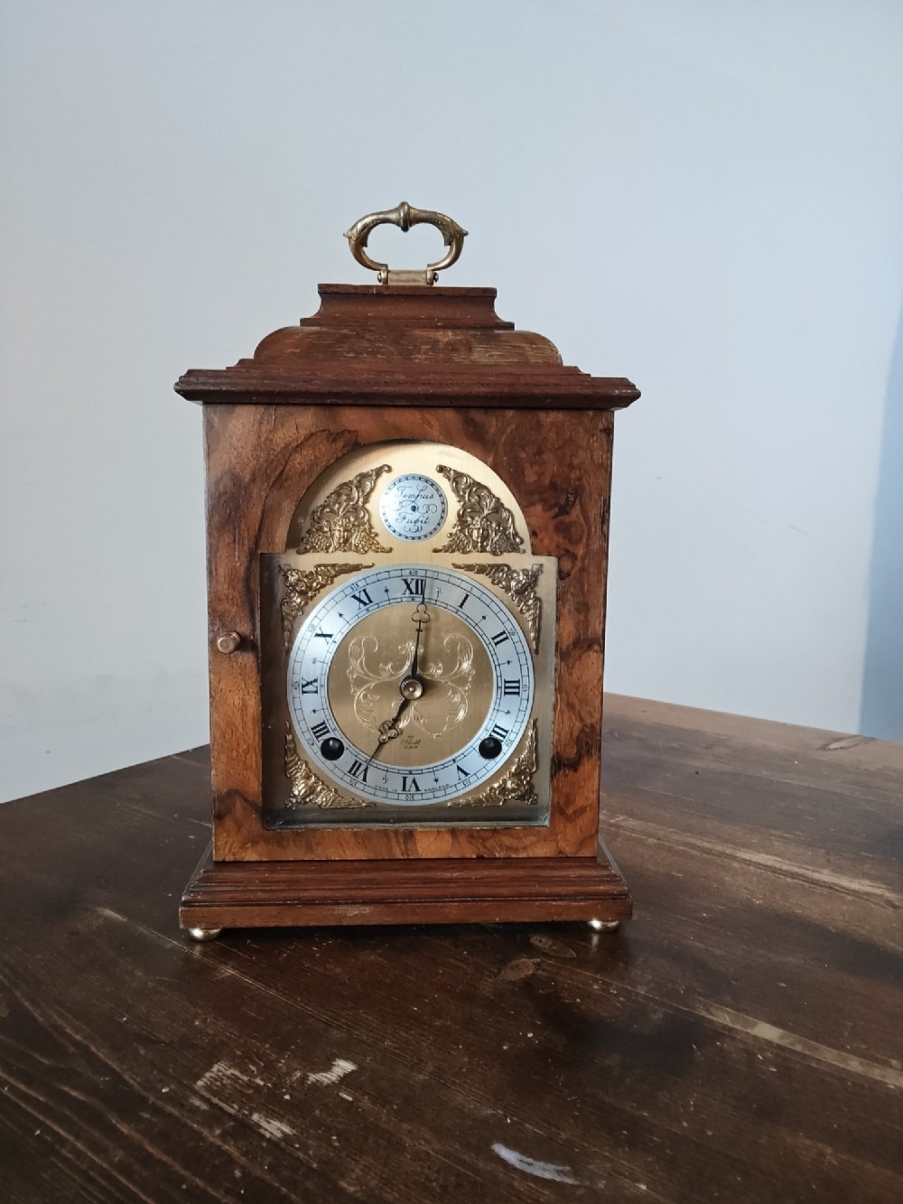 A rectangular burr walnut bracket clock featuring a top brass handle, arched brass dial face with Roman numerals, and a silvered chapter ring.