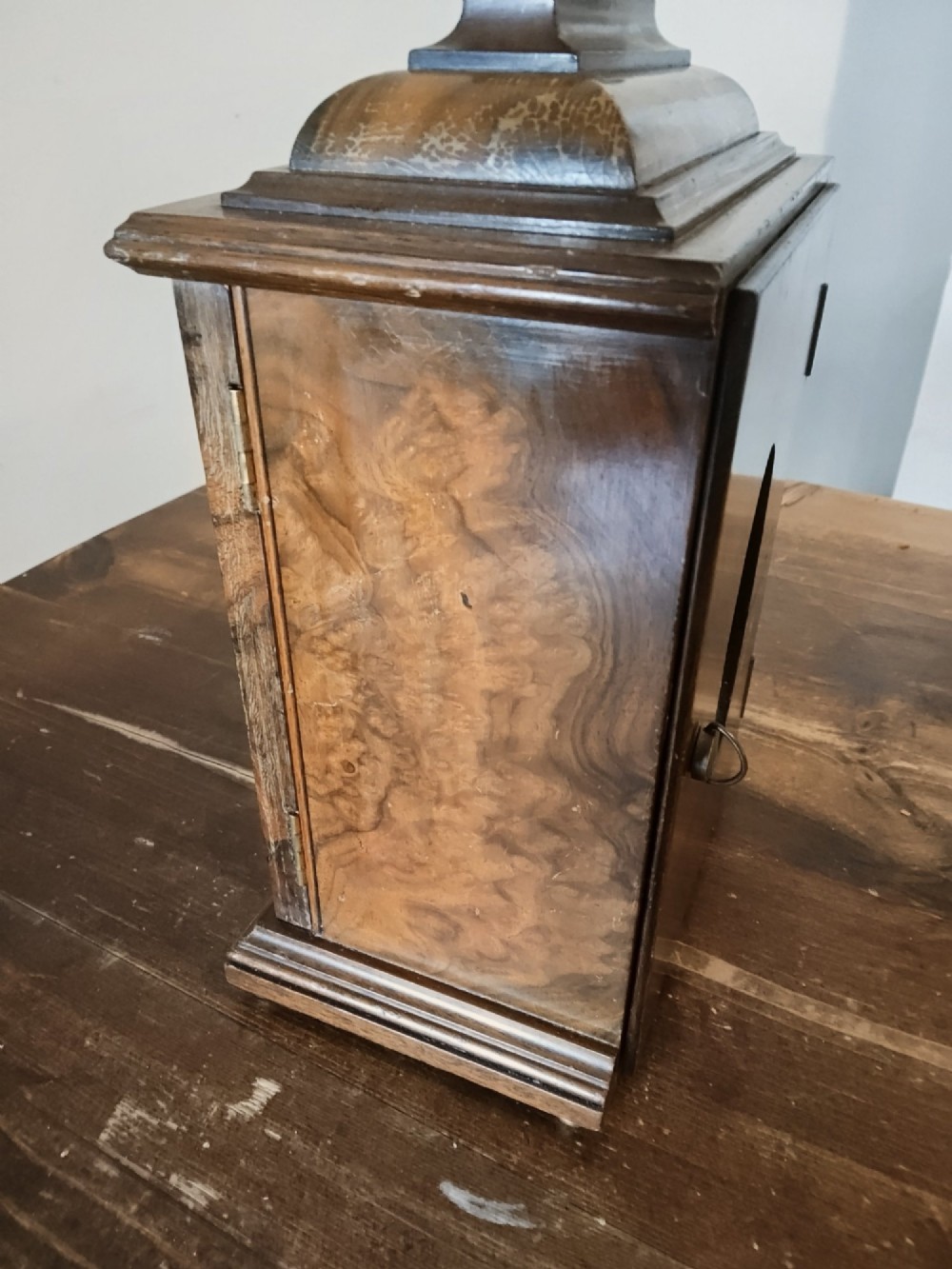 A rectangular burr walnut bracket clock featuring a top brass handle, arched brass dial face with Roman numerals, and a silvered chapter ring. View VI