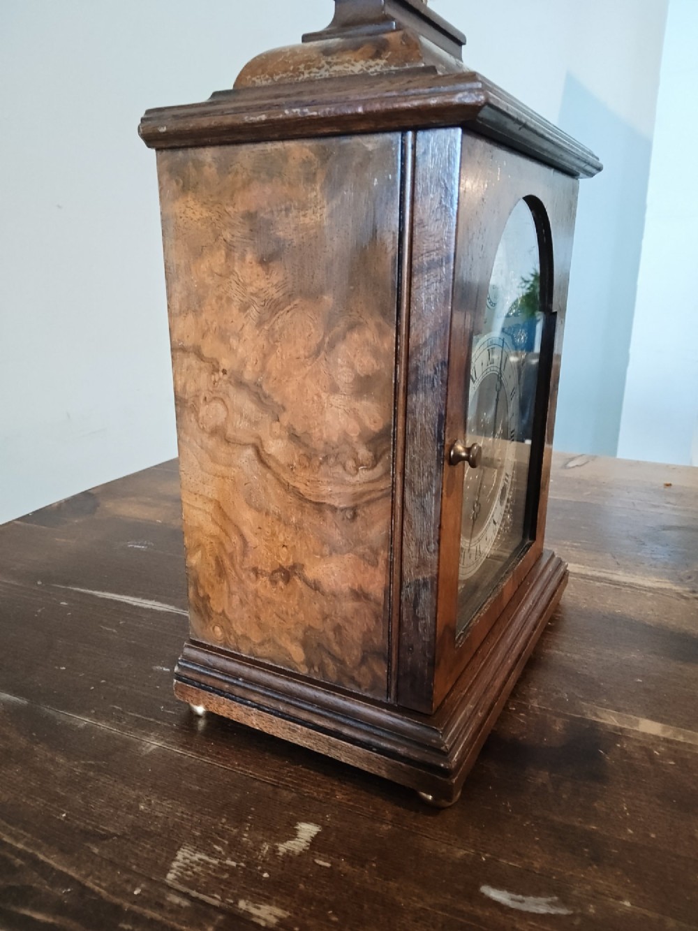A rectangular burr walnut bracket clock featuring a top brass handle, arched brass dial face with Roman numerals, and a silvered chapter ring. View VII