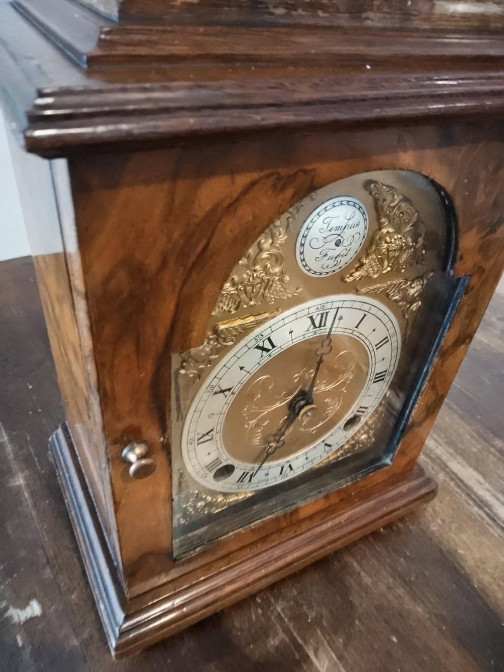 A rectangular burr walnut bracket clock featuring a top brass handle, arched brass dial face with Roman numerals, and a silvered chapter ring. View VIII