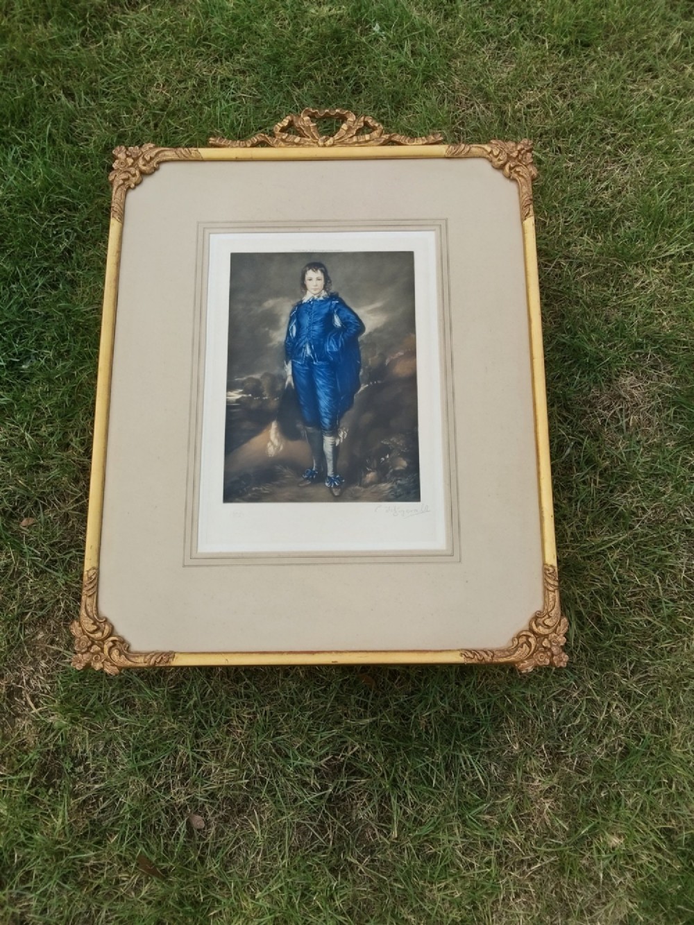 A framed colour portrait print of Thomas Gainsboroughs The Blue Boy, set within a wide beige mount and a gold giltwood frame topped with a bow cresting.