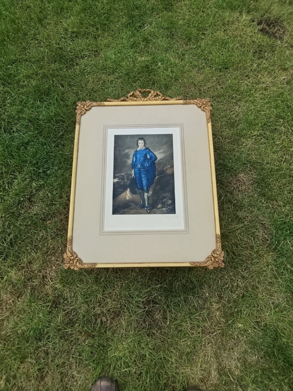 A framed colour portrait print of Thomas Gainsboroughs The Blue Boy, set within a wide beige mount and a gold giltwood frame topped with a bow cresting. View II