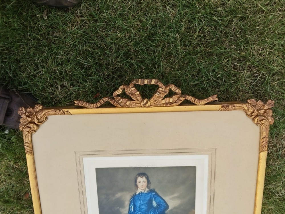 A framed colour portrait print of Thomas Gainsboroughs The Blue Boy, set within a wide beige mount and a gold giltwood frame topped with a bow cresting. View V