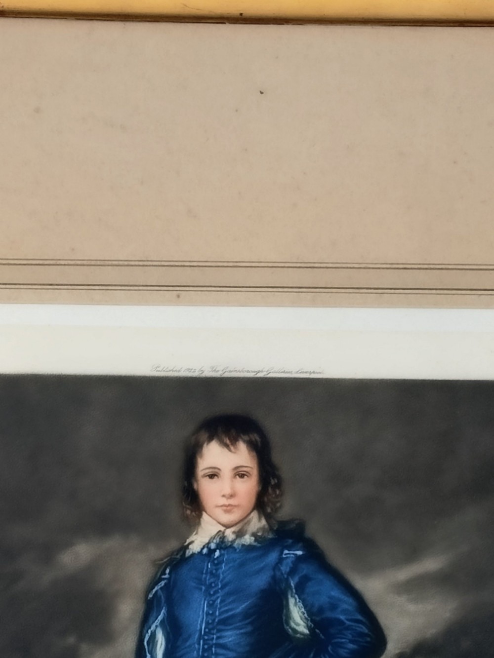 A framed colour portrait print of Thomas Gainsboroughs The Blue Boy, set within a wide beige mount and a gold giltwood frame topped with a bow cresting. View X