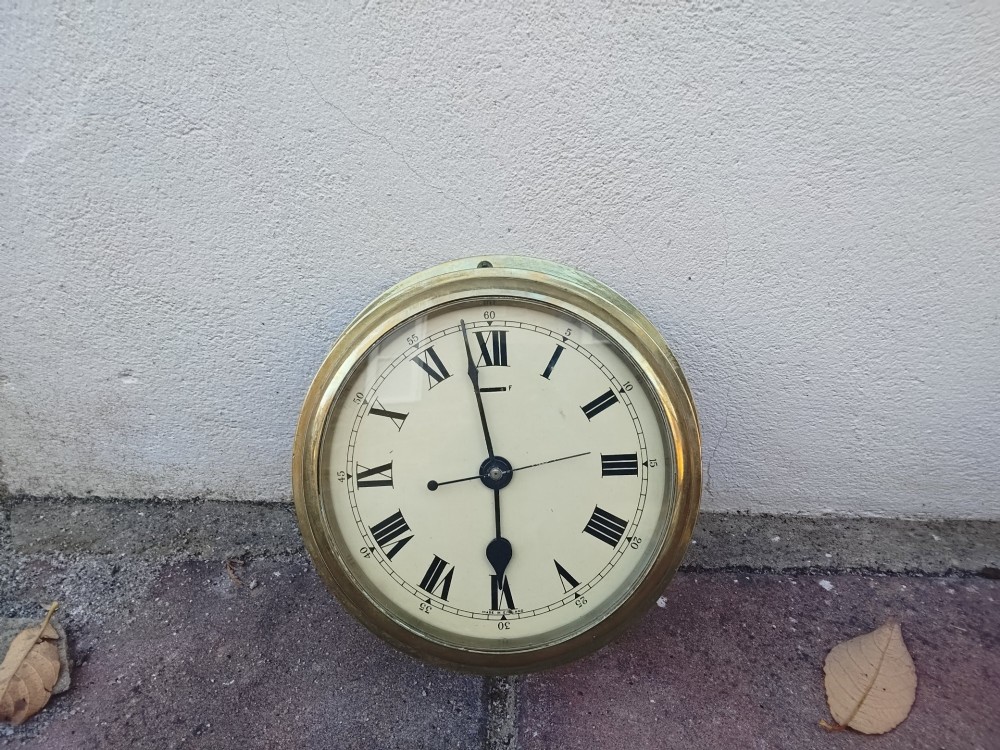 A circular heavy brass marine wall clock featuring a cream dial with black Roman numerals and a flange for wall mounting.