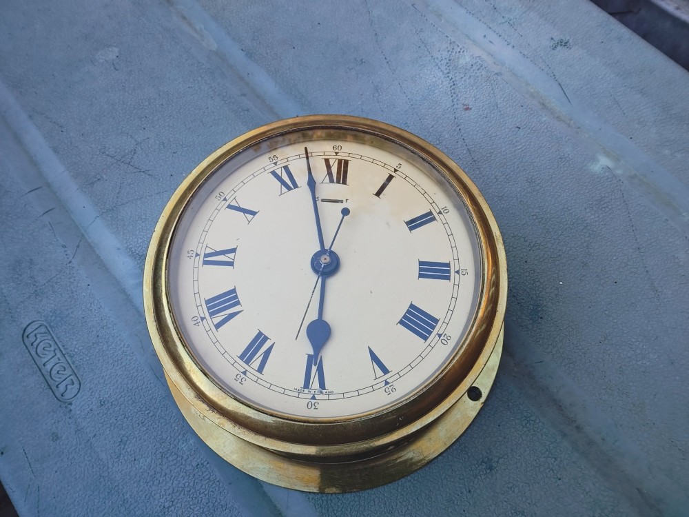 A circular heavy brass marine wall clock featuring a cream dial with black Roman numerals and a flange for wall mounting. View III