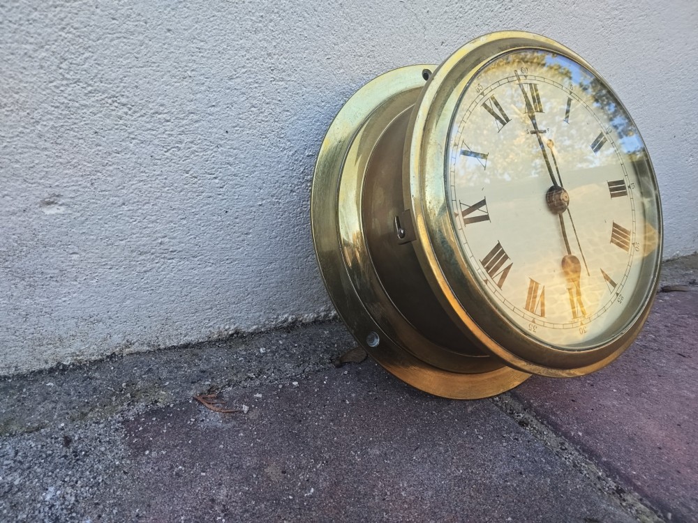A circular heavy brass marine wall clock featuring a cream dial with black Roman numerals and a flange for wall mounting. View IV