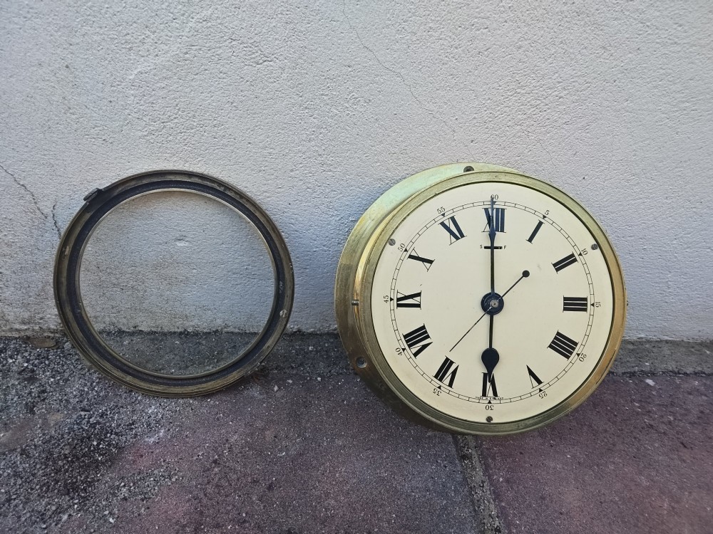 A circular heavy brass marine wall clock featuring a cream dial with black Roman numerals and a flange for wall mounting. View VIII