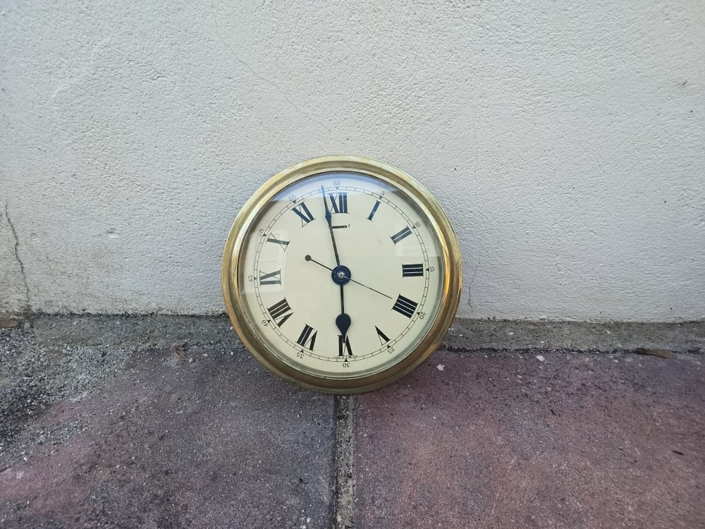 A circular heavy brass marine wall clock featuring a cream dial with black Roman numerals and a flange for wall mounting. View XI