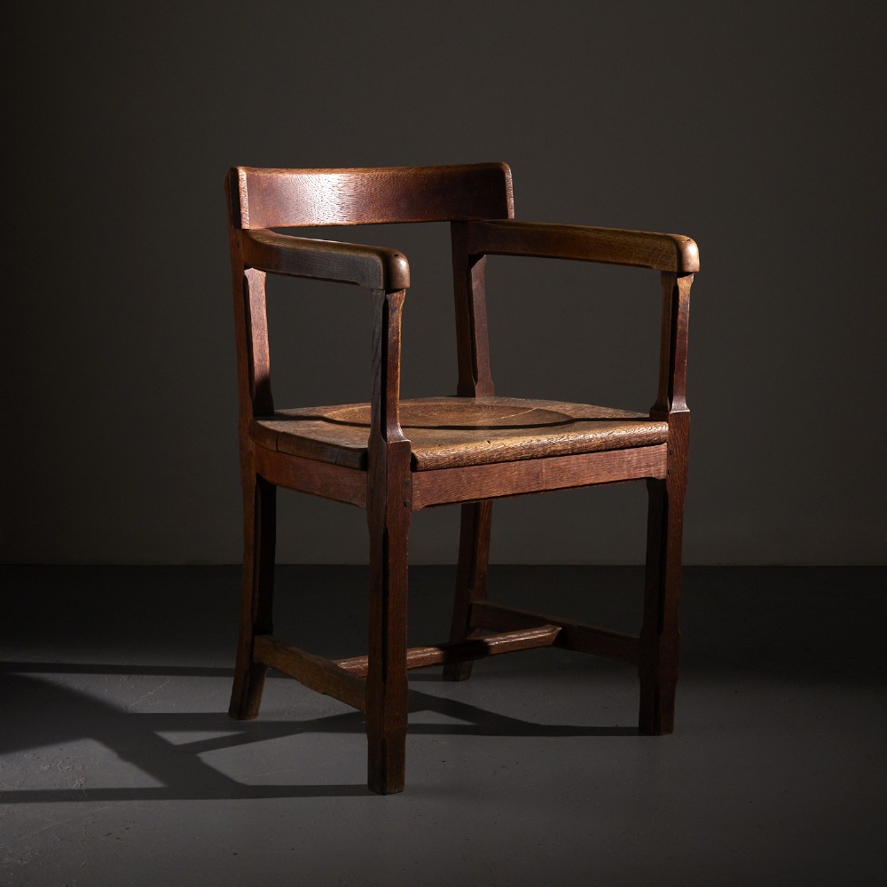 A solid oak Arts and Crafts armchair with flat armrests, a curved backrest, and a contoured wooden seat.