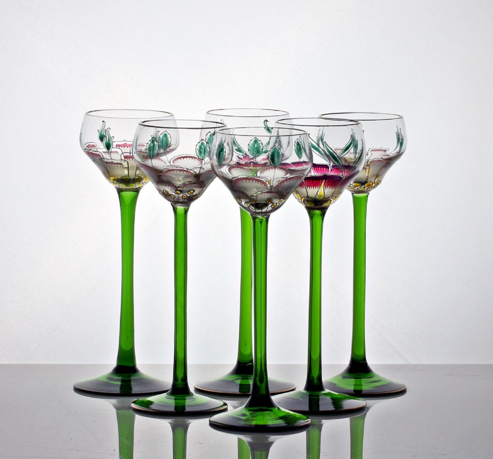 A set of six Art Nouveau style cordial glasses featuring tall, slender green stems and clear bowls decorated with hand-painted enamel floral motifs in pink, green, and white.