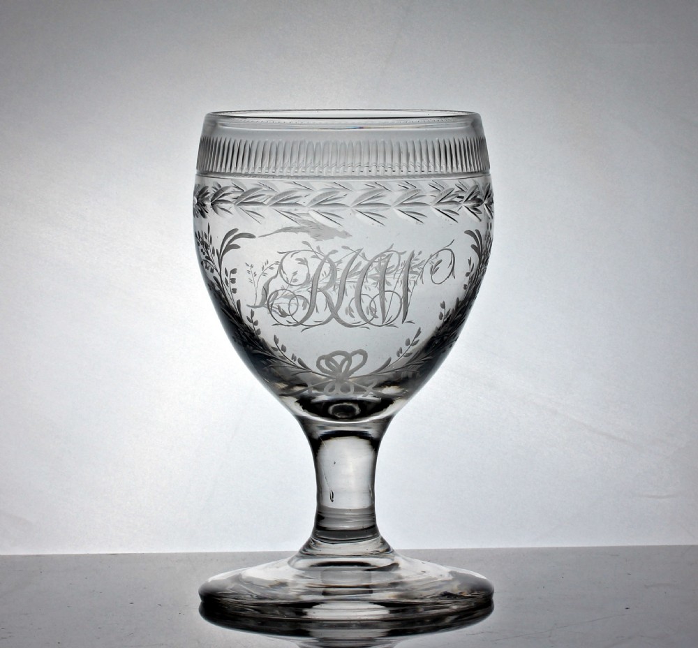 A clear Georgian glass goblet featuring an ovoid bowl with a wheel-engraved monogram and floral wreath above a short plain stem and circular foot.