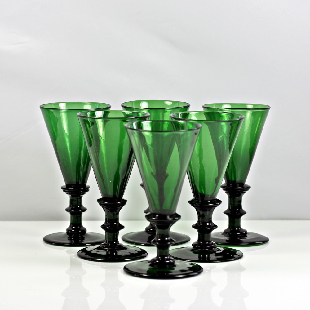 A set of six dark green hand-blown glass wine glasses featuring conical bowls and knopped stems arranged on a reflective white surface.