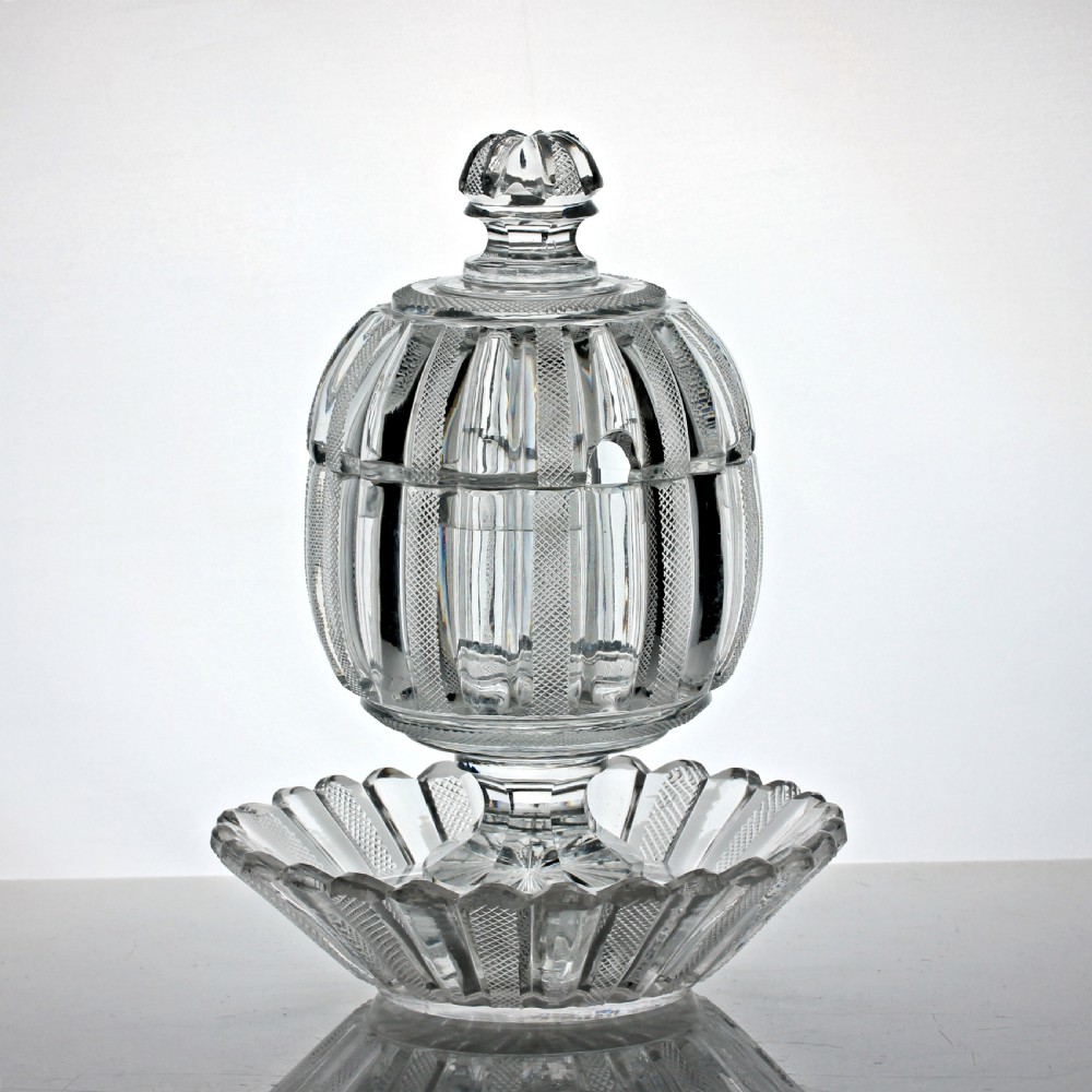 A clear cut glass preserve pot with a fitted lid and knopped finial resting on a matching scalloped glass saucer base.