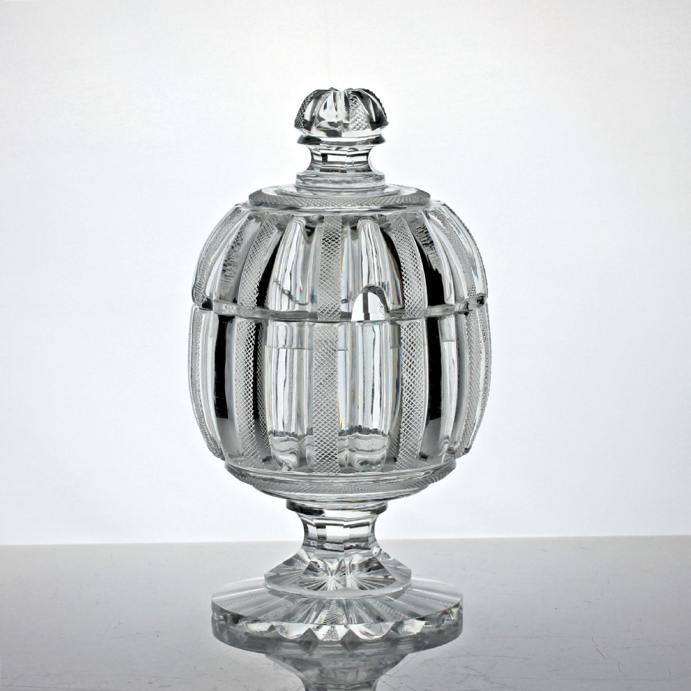 A clear cut glass preserve pot with a fitted lid and knopped finial resting on a matching scalloped glass saucer base. View II