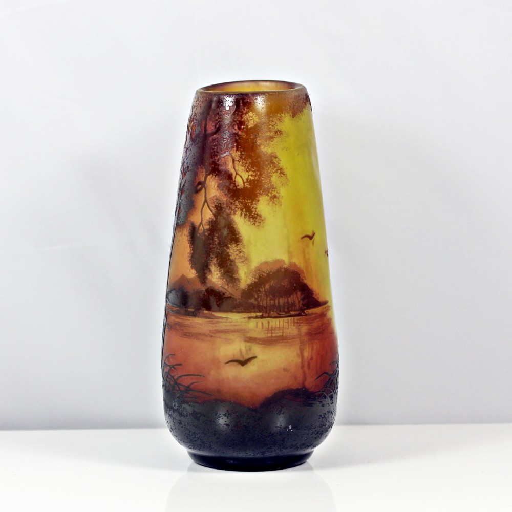 An Art Nouveau cameo glass vase featuring an acid-etched sunset landscape scene with trees, water, and birds in shades of amber, yellow, and dark brown.
