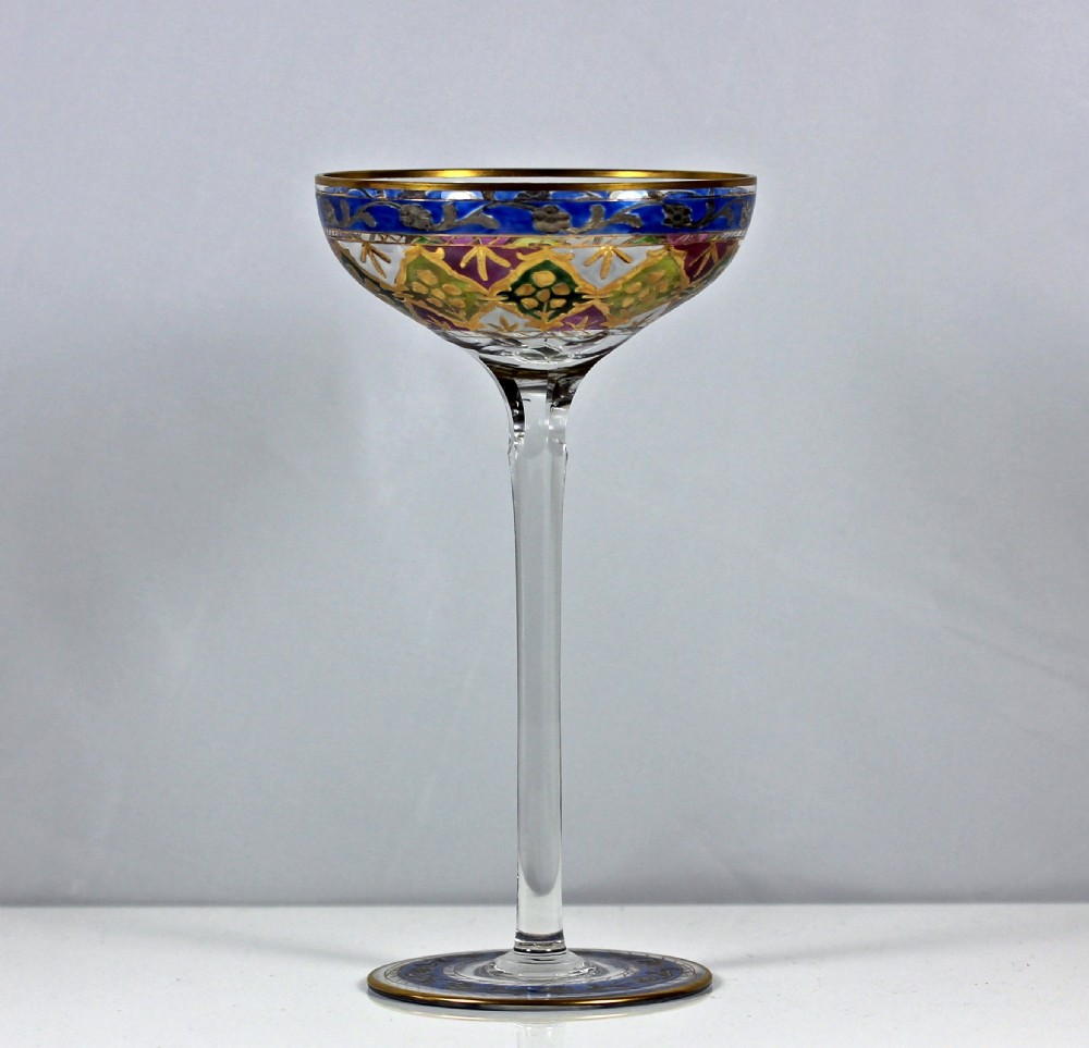 A clear glass champagne coupe with a tall slender stem, decorated with colourful geometric enamel patterns and gold gilding around the rim and foot.