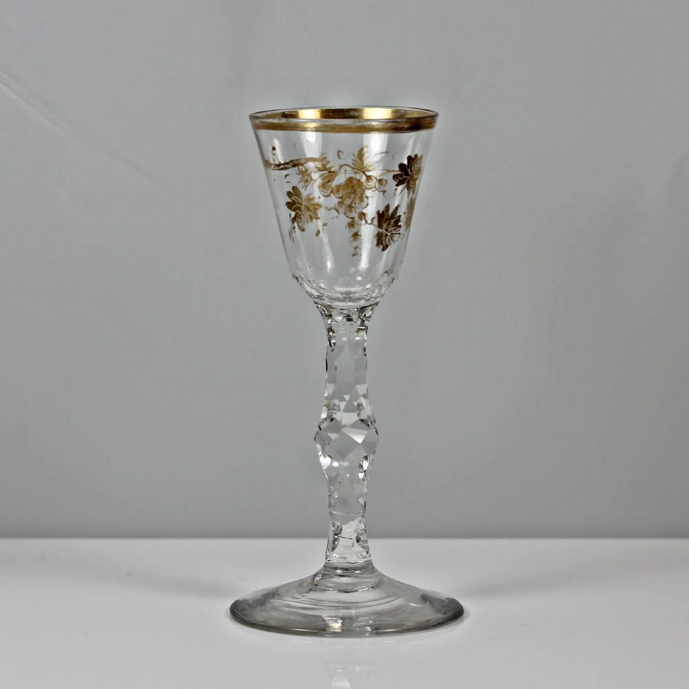 An 18th-century clear lead glass wine glass featuring a gilded vine leaf motif on the bowl and a geometric facet cut stem.