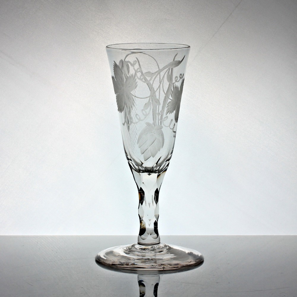 A tall Georgian clear glass ale flute featuring engraved hop and barley motifs on the bowl and a hexagonal facet-cut stem.