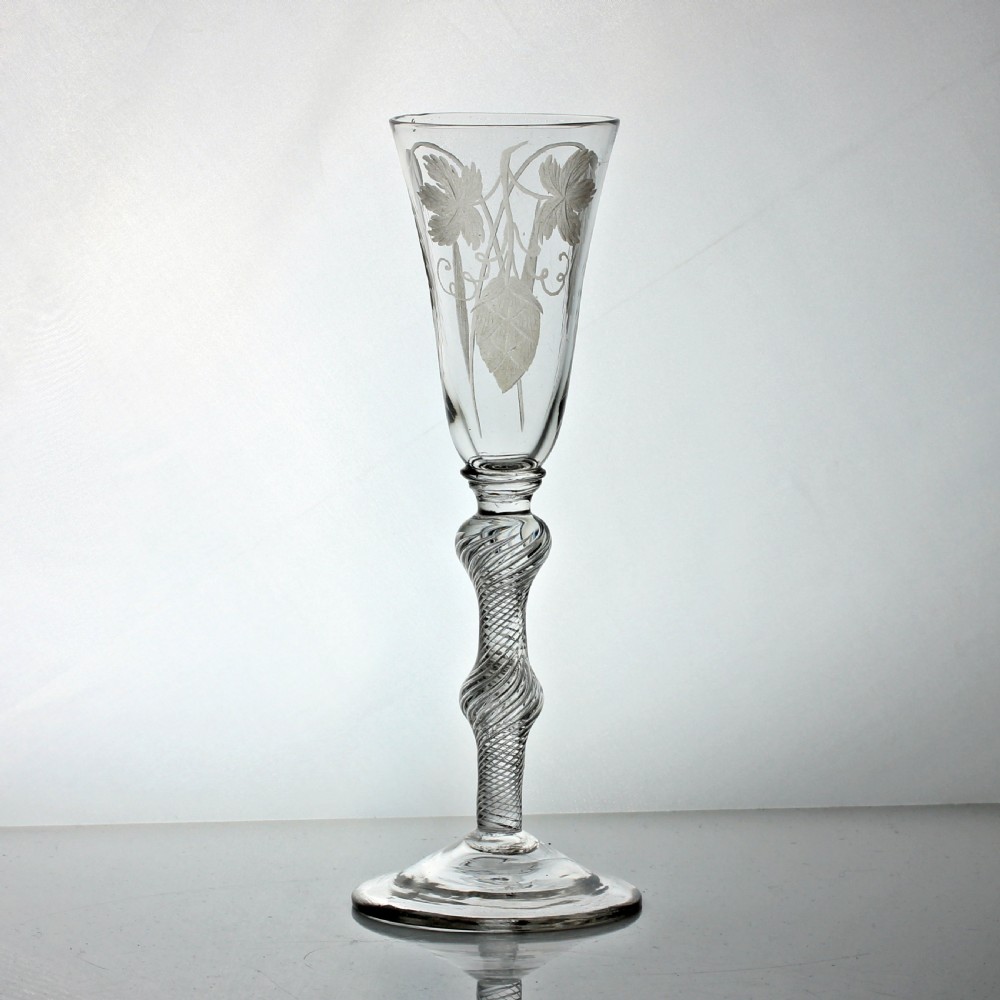A tall clear glass ale flute with wheel-engraved hop and barley motifs above a knopped multi-spiral air twist stem and conical foot.