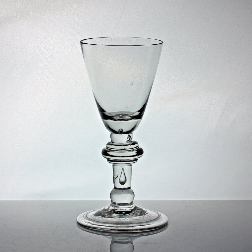 An antique clear glass wine goblet featuring a conical bowl, a stem with a triple annular knop containing an air bubble tear, resting on a wide circular foot.
