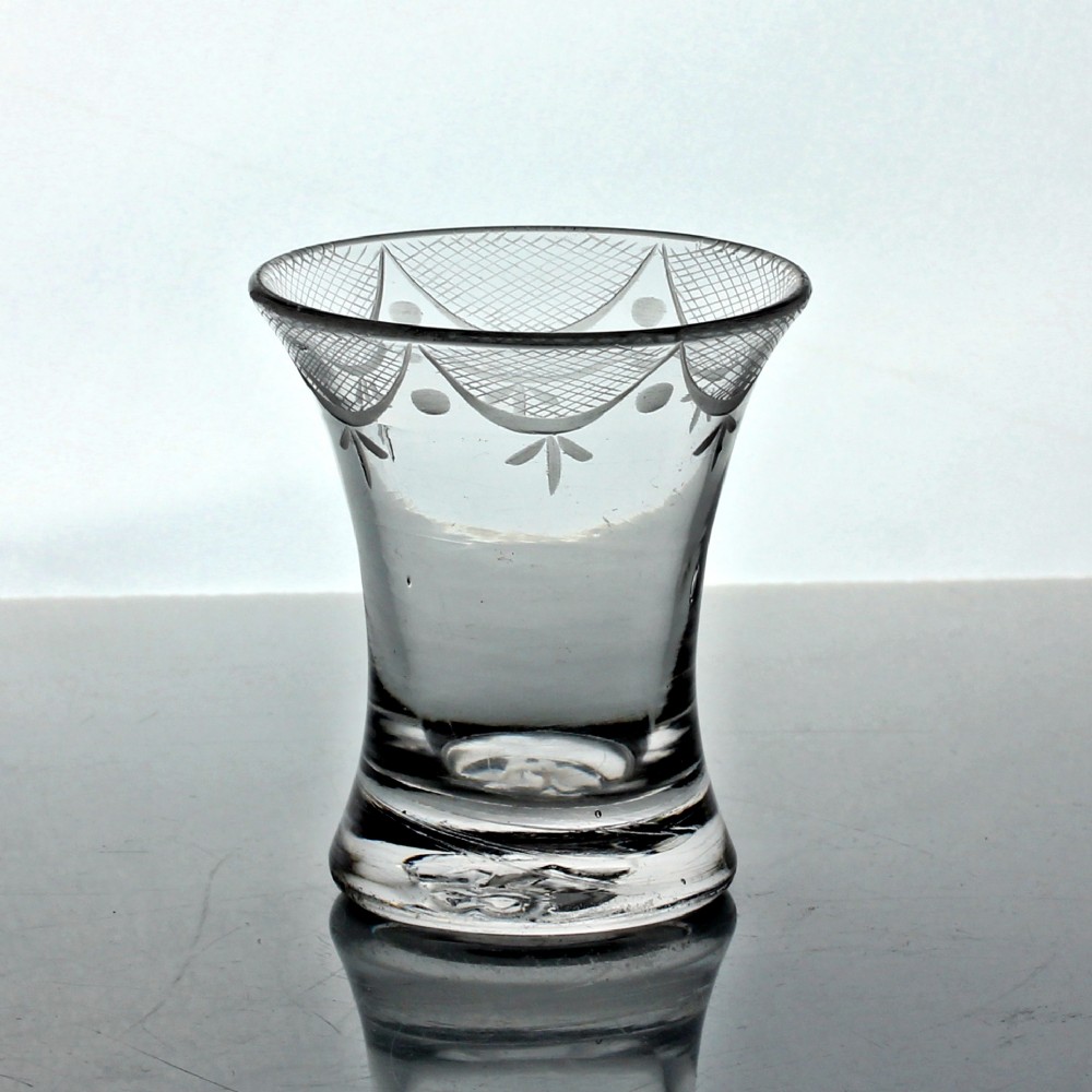 A small clear Georgian firing glass featuring a flared rim, heavy circular base, and wheel-engraved swag and tail decoration.