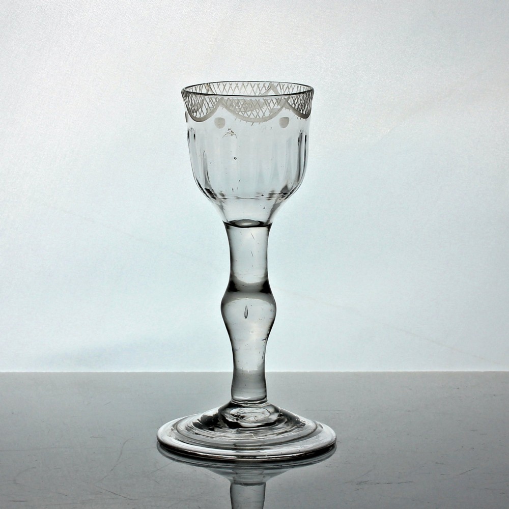 An 18th-century clear lead glass wine goblet featuring a swag and tail engraved rim, a knopped balustroid stem, and a flat circular foot.