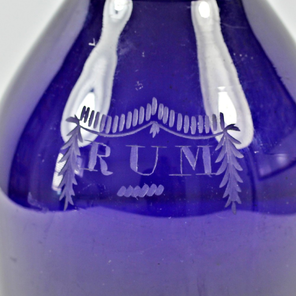 A deep cobalt blue glass decanter featuring three rings around the neck, a matching round stopper, and the word RUM etched within a decorative swag on the front. View II