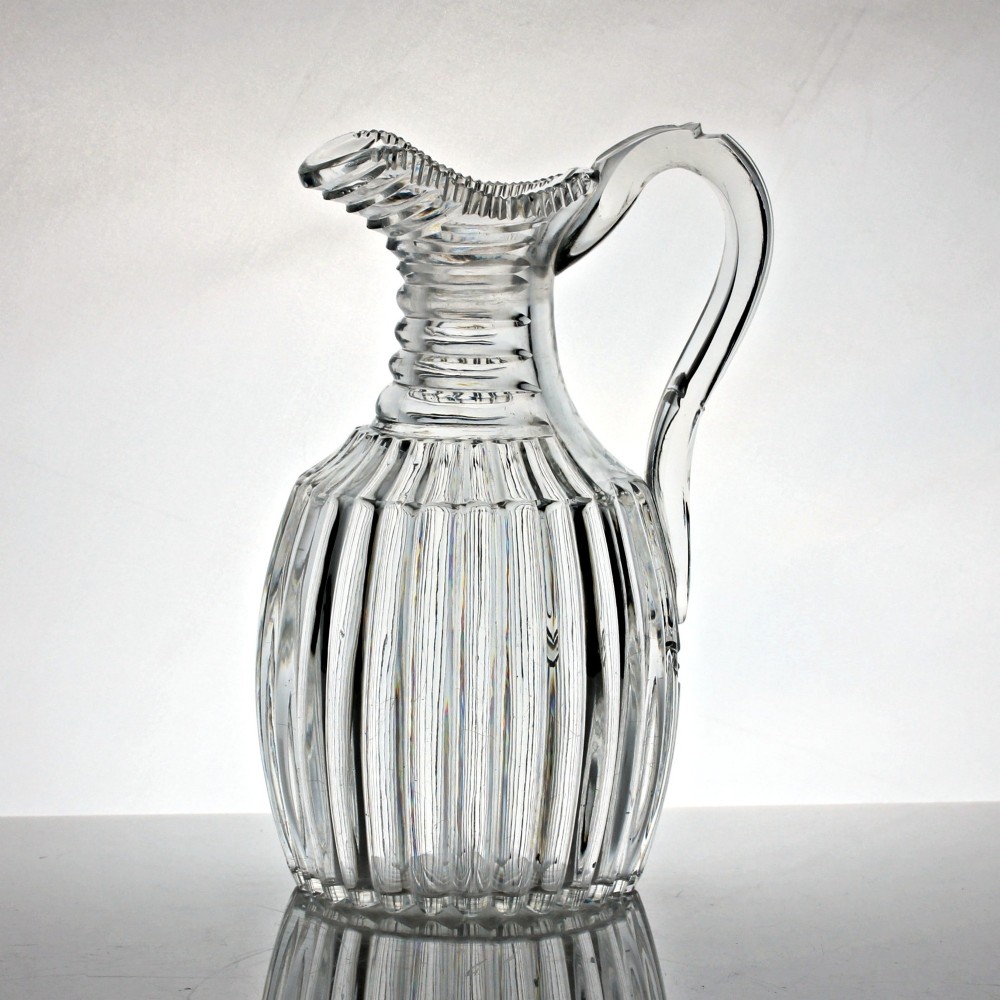 A clear Georgian lead crystal water jug featuring vertical pillar cutting around the lower body, neck rings, and a notched poured spout with an applied handle.