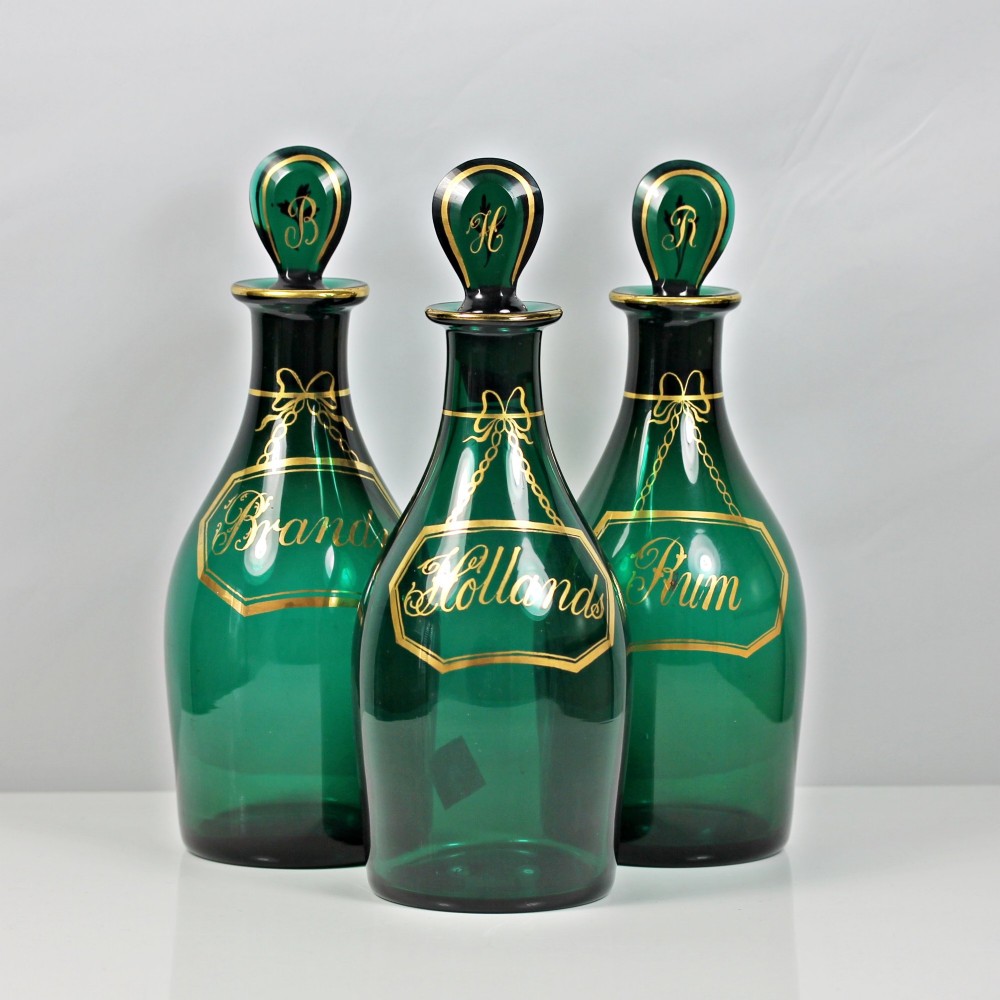 A set of three Georgian Bristol green glass decanters with gilded labels for Brandy, Hollands, and Rum, featuring monograms on the disc stoppers.