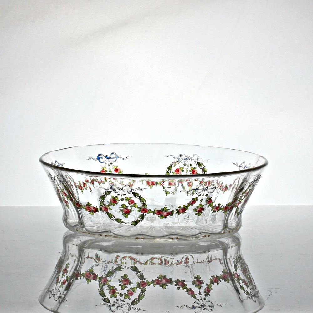 An oval clear glass bowl featuring hand-painted enamelled garlands of red roses, green foliage, and blue ribbons along the exterior with a gilded rim.