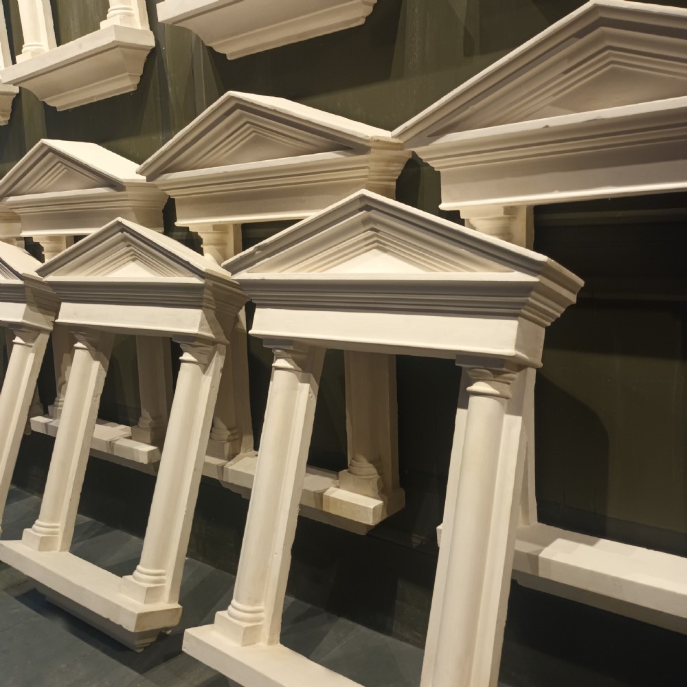 A white plaster tabernacle picture frame featuring a triangular pediment supported by two smooth classical columns resting on a projecting plinth base. View II