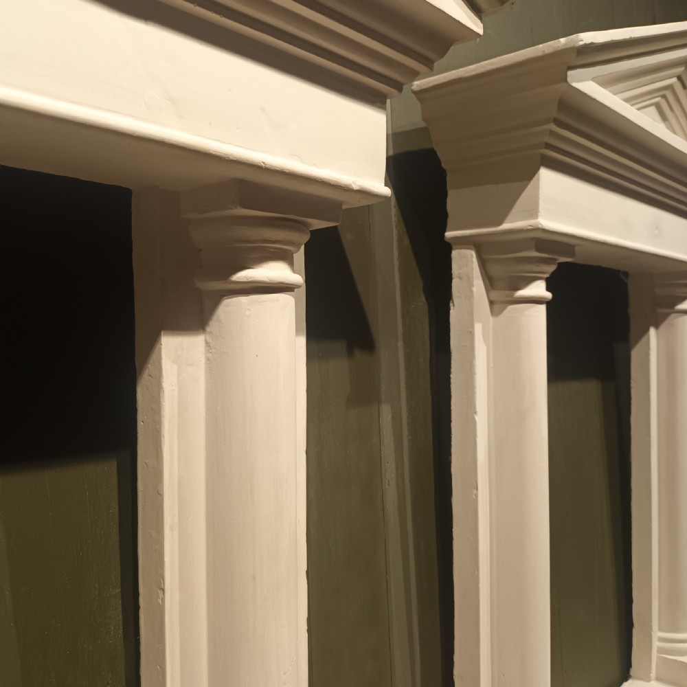 A white plaster tabernacle picture frame featuring a triangular pediment supported by two smooth classical columns resting on a projecting plinth base. View VII