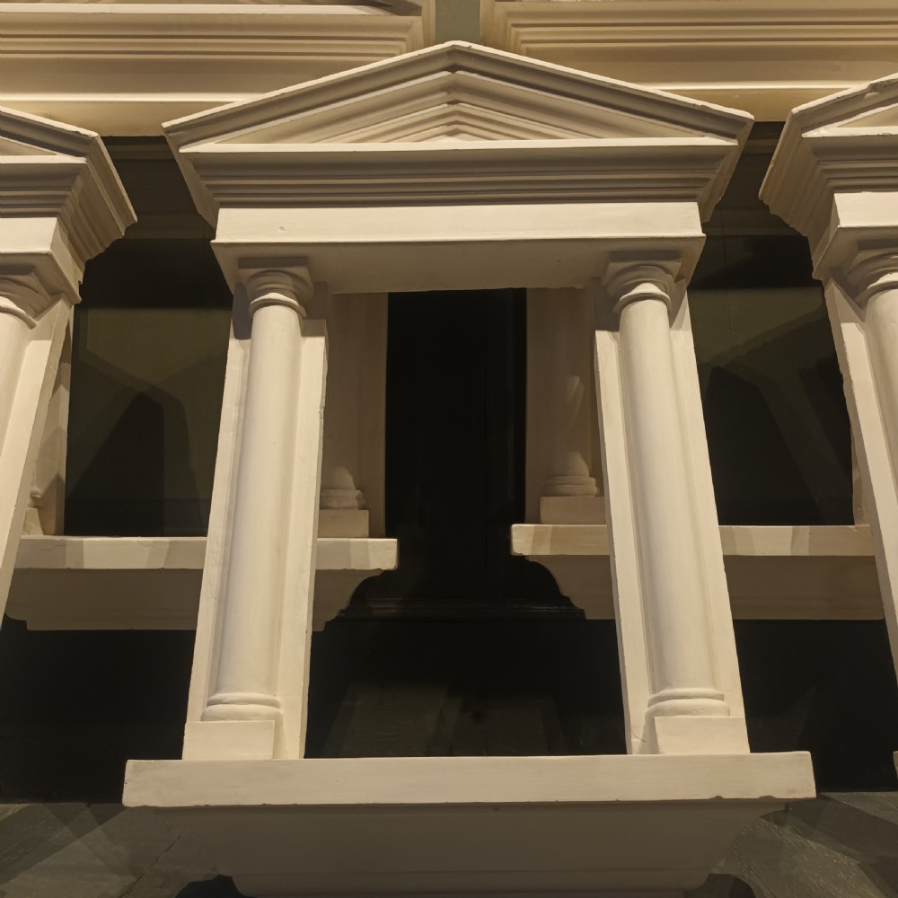 A white plaster tabernacle picture frame featuring a triangular pediment supported by two smooth classical columns resting on a projecting plinth base. View IX