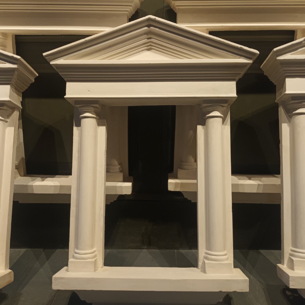 A white plaster tabernacle picture frame featuring a triangular pediment supported by two smooth classical columns resting on a projecting plinth base. View XII