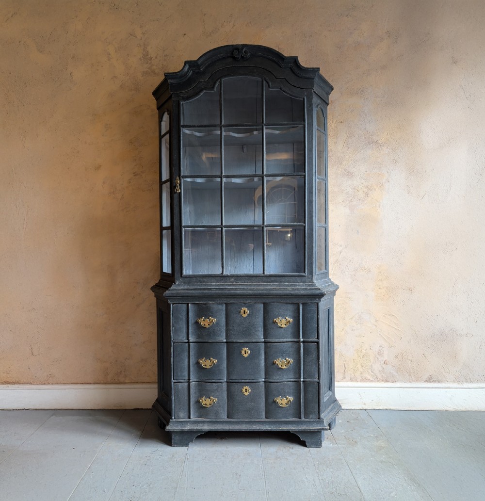 A tall dark grey painted Dutch vitrine cabinet featuring a shaped top hood, glazed upper doors, and three lower drawers with brass handles.