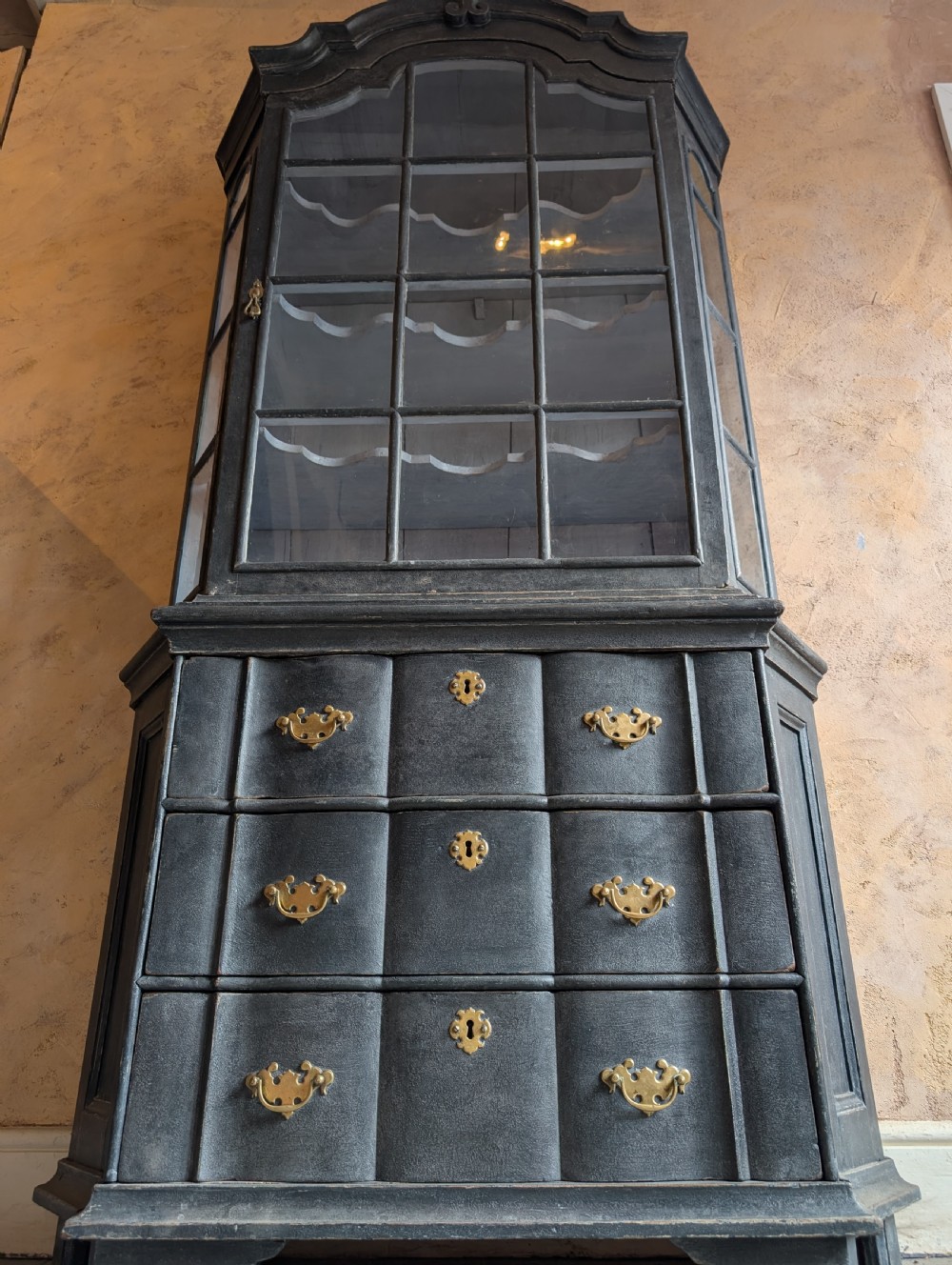 A tall dark grey painted Dutch vitrine cabinet featuring a shaped top hood, glazed upper doors, and three lower drawers with brass handles. View III