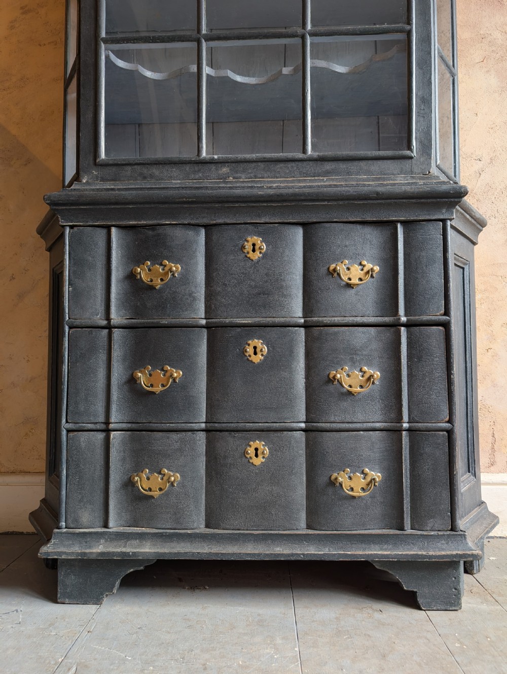 A tall dark grey painted Dutch vitrine cabinet featuring a shaped top hood, glazed upper doors, and three lower drawers with brass handles. View V