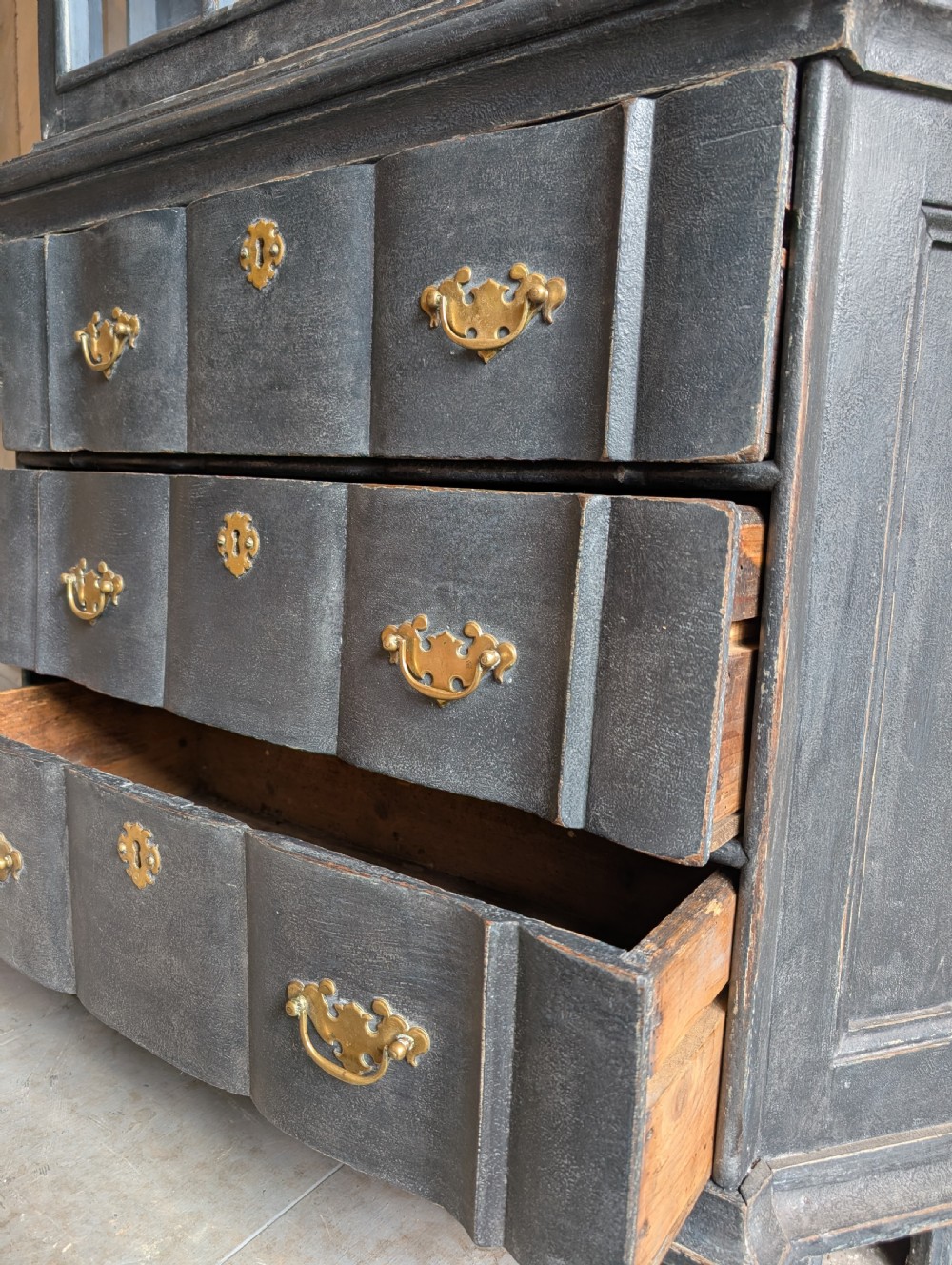 A tall dark grey painted Dutch vitrine cabinet featuring a shaped top hood, glazed upper doors, and three lower drawers with brass handles. View VII