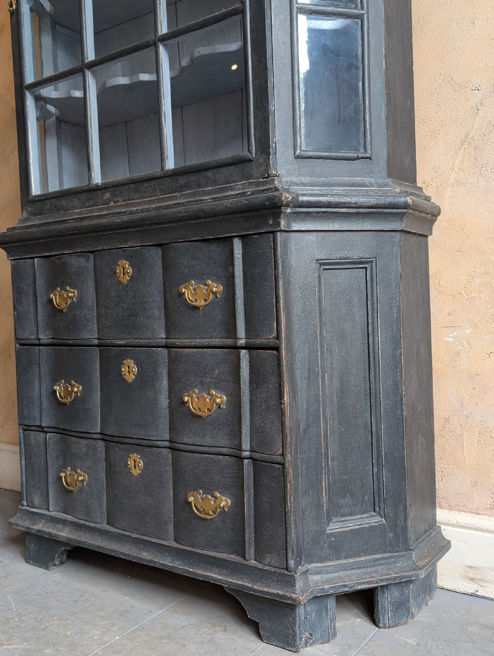 A tall dark grey painted Dutch vitrine cabinet featuring a shaped top hood, glazed upper doors, and three lower drawers with brass handles. View IX