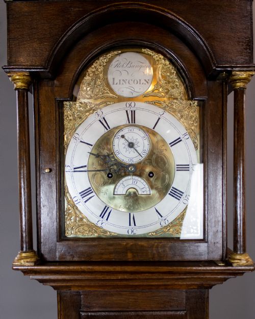 fig 1. An antique dark oak longcase clock featuring an arched brass dial with Roman numerals and a pediment hood.
