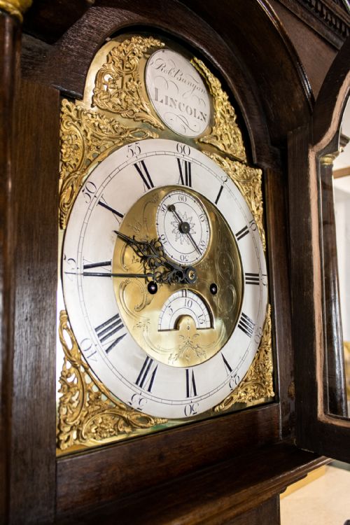 fig 2. An antique dark oak longcase clock featuring an arched brass dial with Roman numerals and a pediment hood.