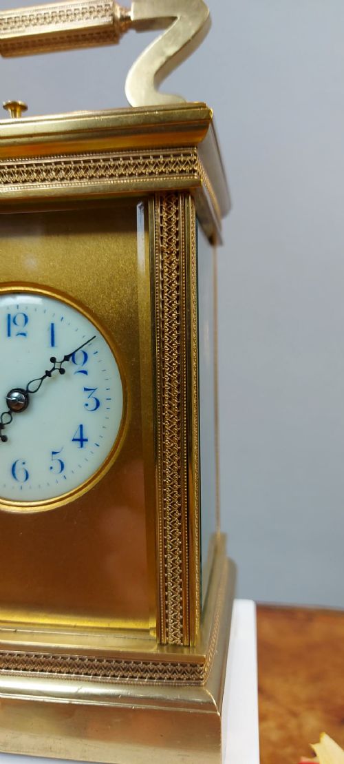 fig 1. A rectangular gilt brass repeating carriage clock featuring a stylized carrying handle, patterned border trim, and a circular white enamel dial with blue numerals.
