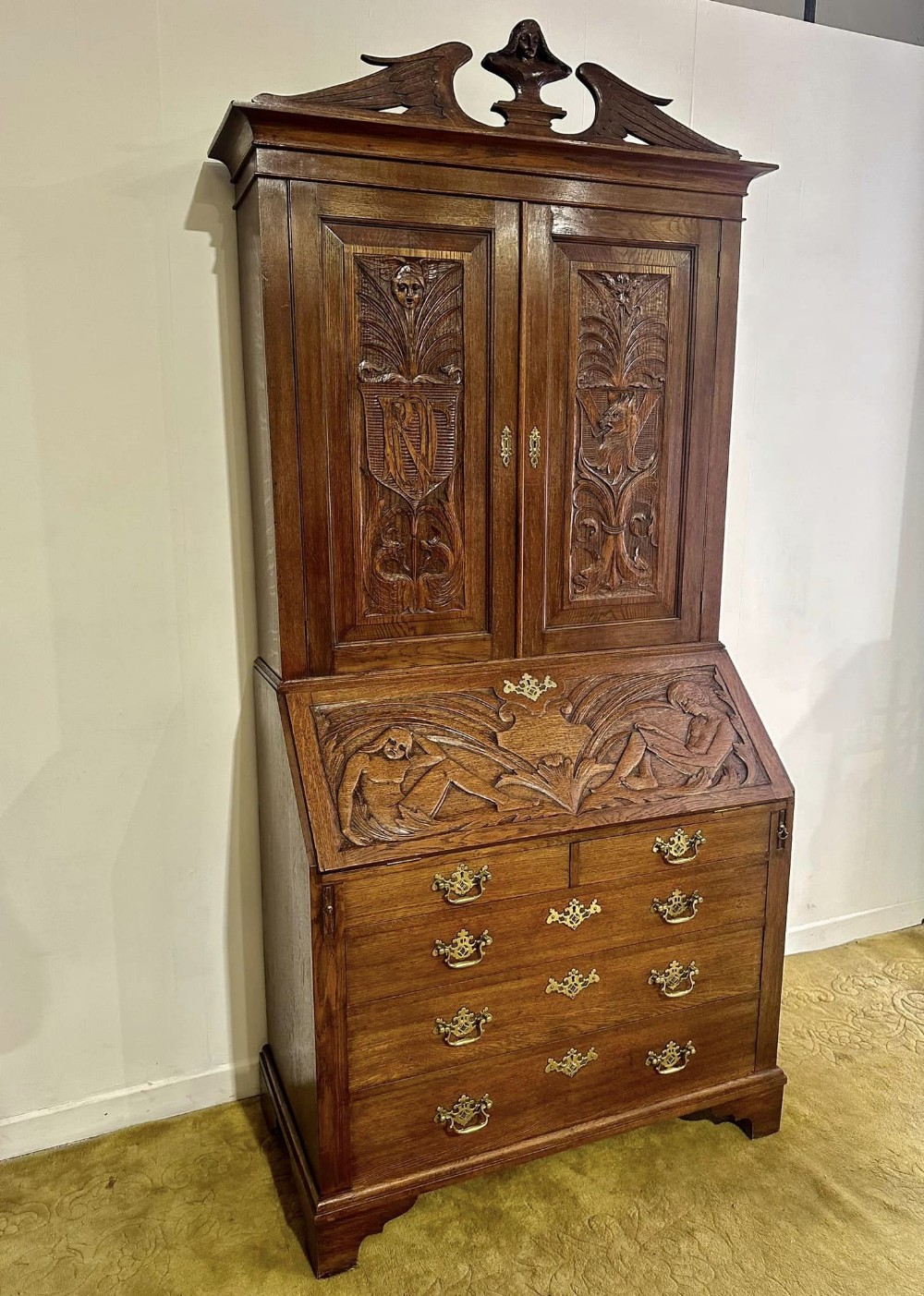 A Victorian carved oak bureau bookcase featuring a scrolled pediment top, two carved upper doors, a slope-front writing surface with relief carvings, and four lower drawers with brass handles.