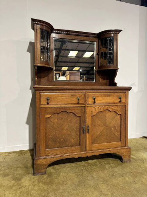 fig 1. A golden oak sideboard featuring a central mirror back flanked by two curved leaded glass cabinets above two drawers and two cupboard doors.